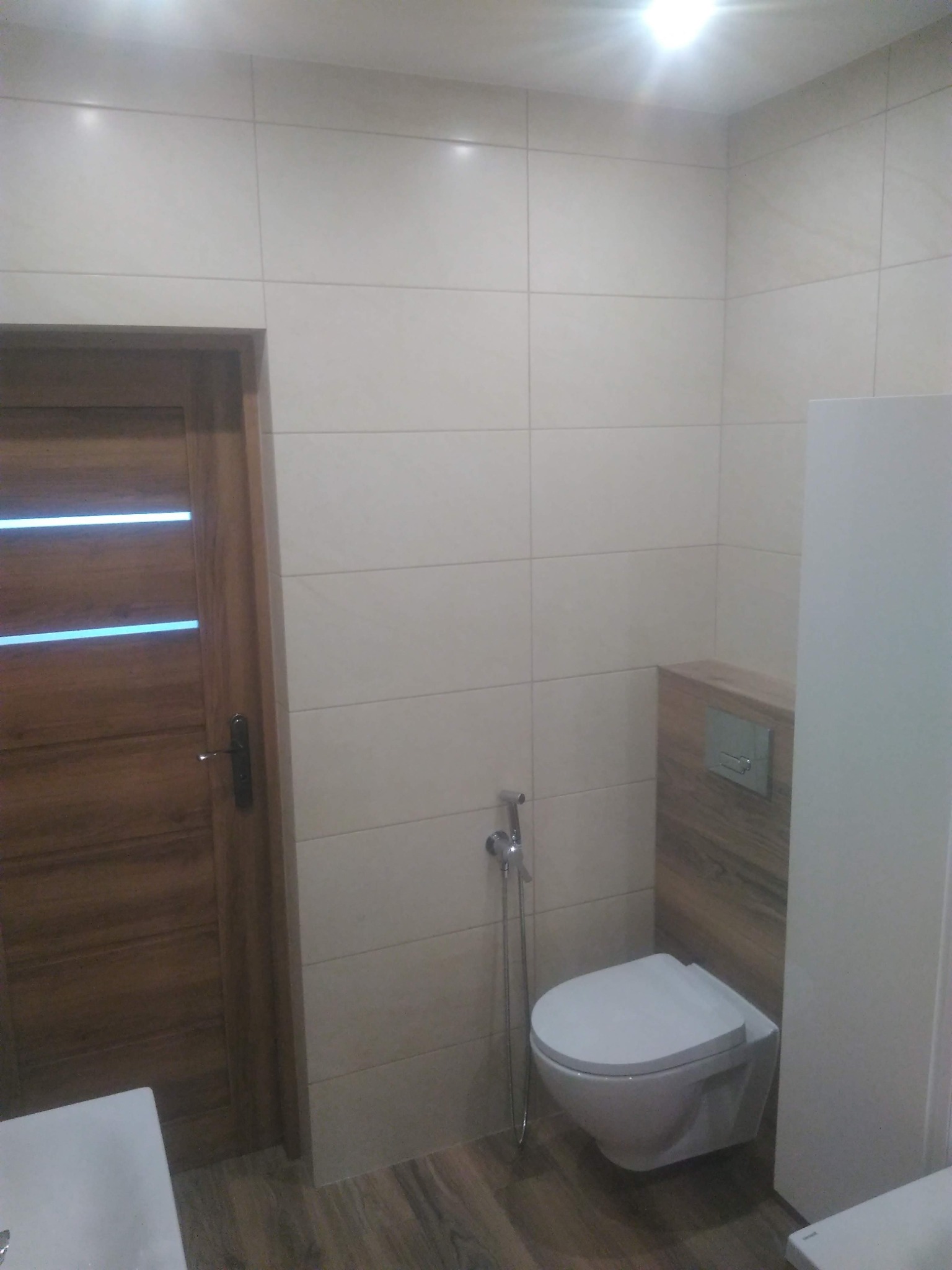 Nowoczesna łazienka z beżowymi płytkami, podwieszaną toaletą z bidetką i drewnopodobną podłogą oraz elementami dekoracyjnymi z motywem drewna.