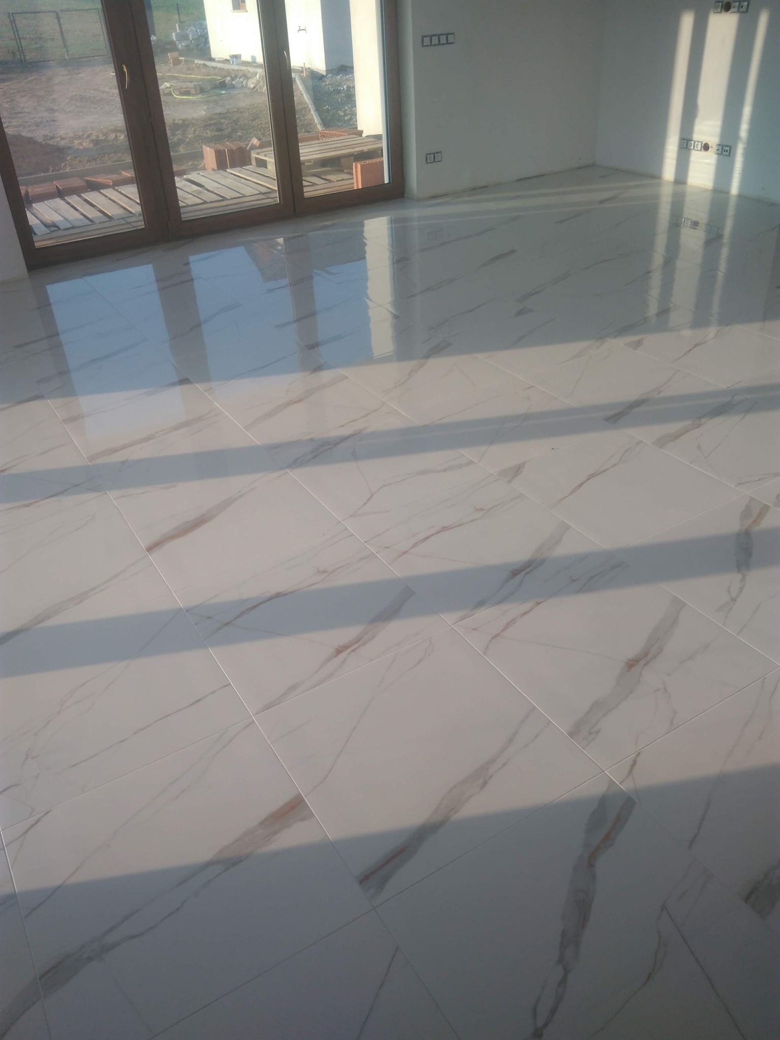 Błyszczące, białe płytki podłogowe imitujące marmur, ułożone w pustym, jasnym pokoju z dużym oknem balkonowym i widocznymi puszkami elektrycznymi na ścianach.