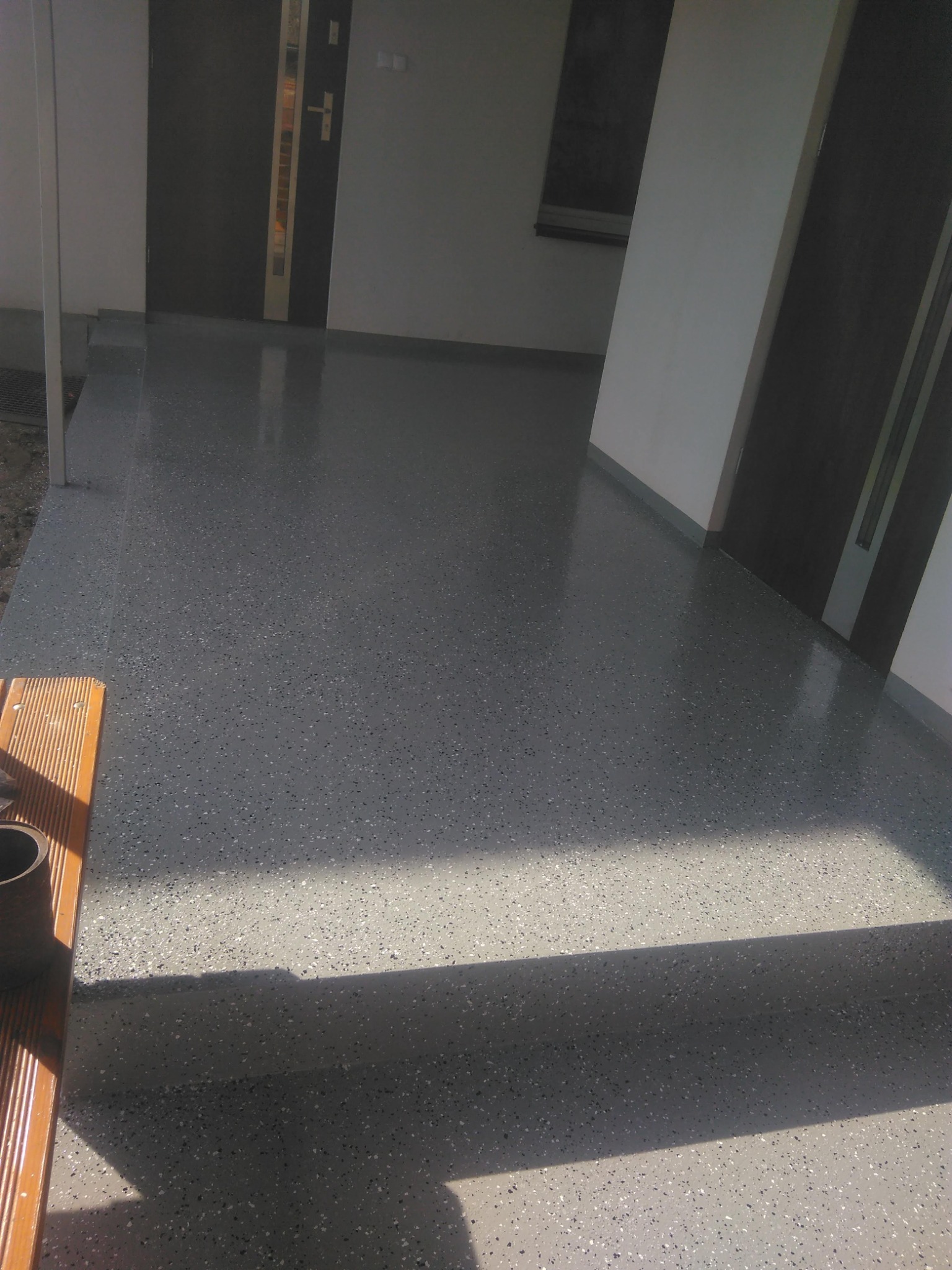 Szara posadzka z drobnymi plamkami, prowadząca do nowoczesnych drzwi wejściowych z metalowymi akcentami, oświetlona naturalnym światłem.