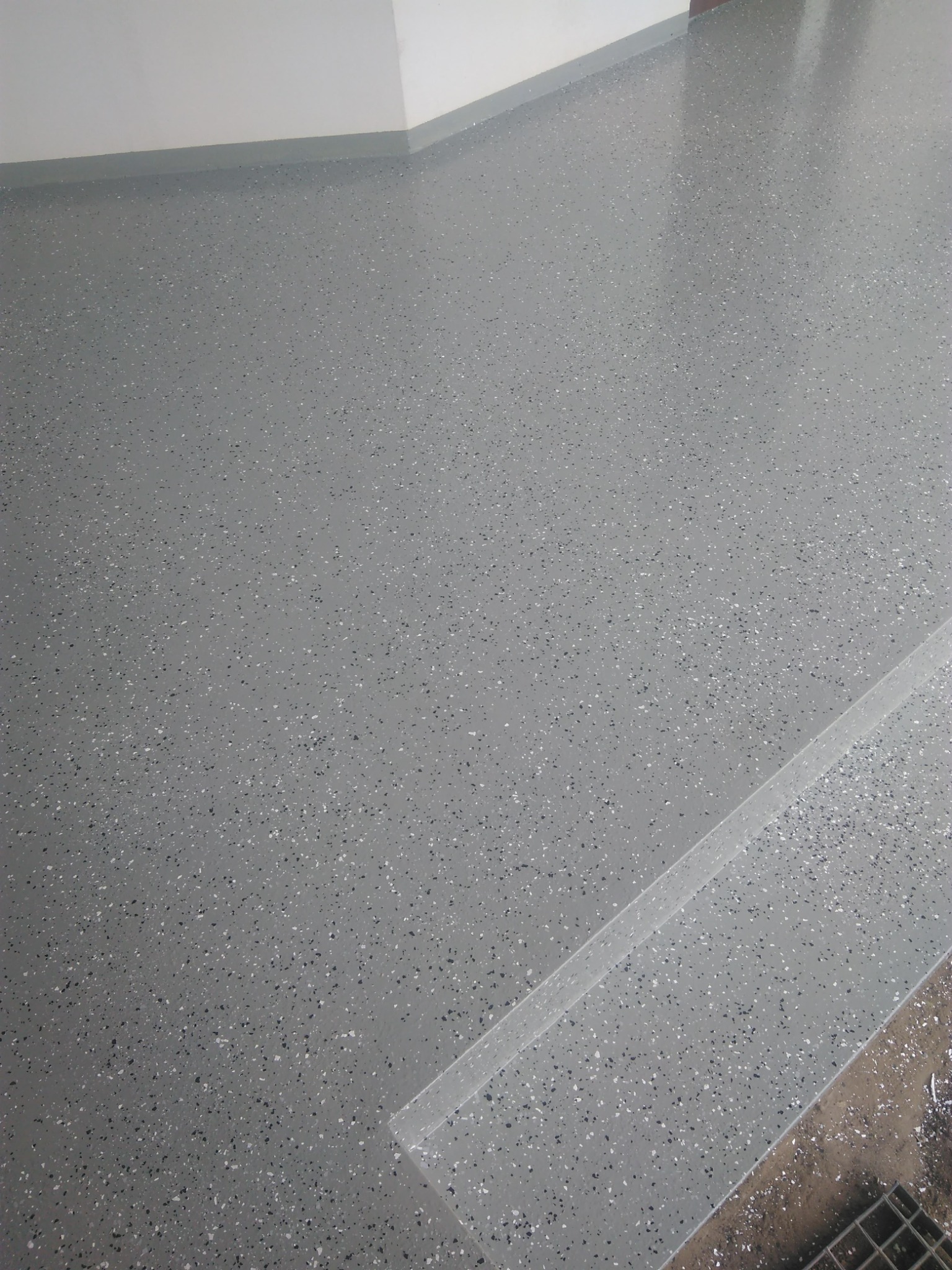 Szara, błyszcząca podłoga z żywicy epoksydowej z czarnymi i białymi drobinkami, widoczna listwa przypodłogowa i fragment metalowej kratki w rogu.