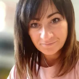 Portret kobiety z brązowymi włosami i delikatnym uśmiechem, prawdopodobnie specjalistki z Płońska, w różowej koszulce z napisem.