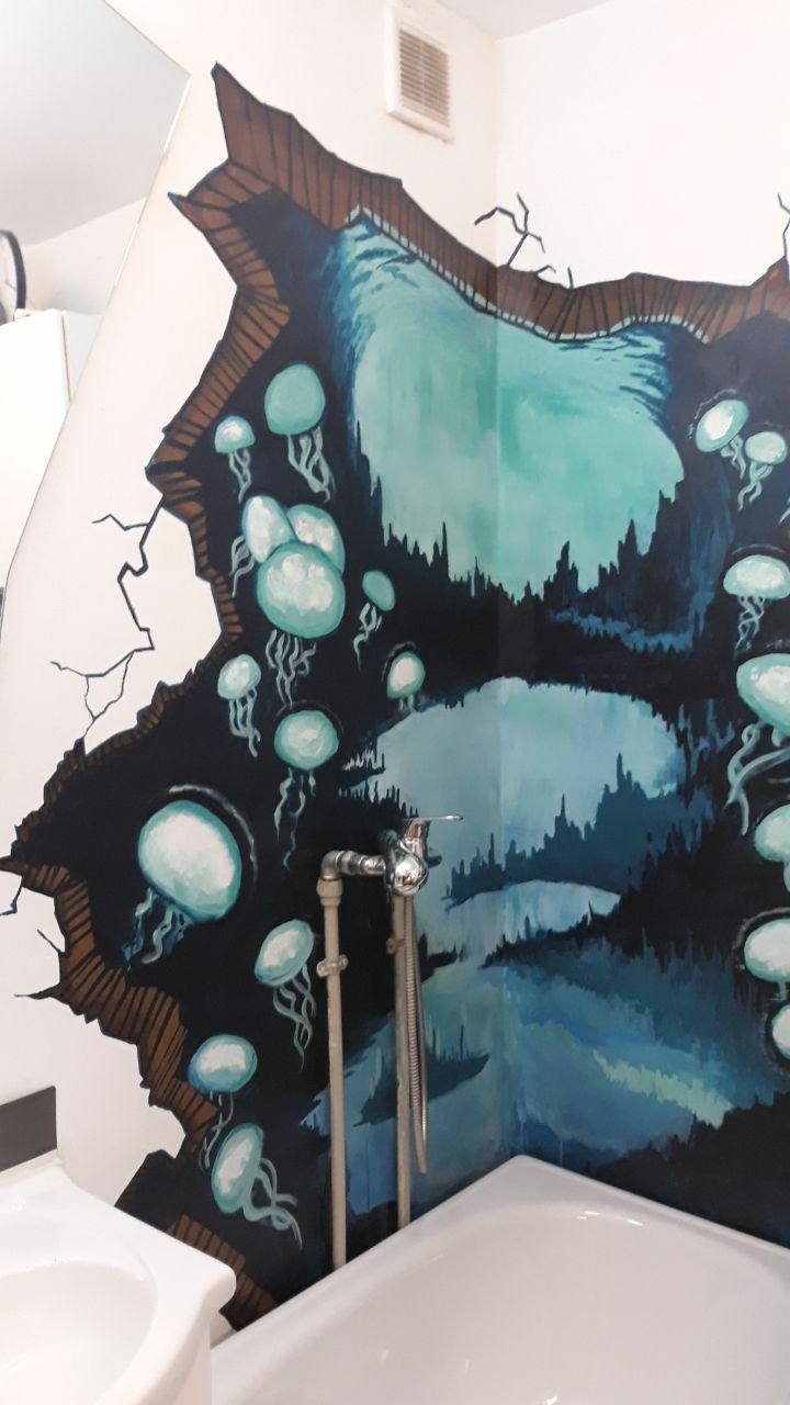 Iluzjonistyczne malarstwo ścienne w łazience przedstawia podwodny krajobraz z meduzami wydobywający się z pękniętej ściany, częściowo zasłonięte przez baterię wannową i umywalkę.