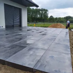 Świeżo ułożony, mokry taras z szarych płyt betonowych przy nowoczesnym domu z szarymi roletami zewnętrznymi, widoczny piasek na obrzeżach i pracownik z zagęszczarką w tle.