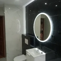 Nowoczesna łazienka z czarno-białymi marmurkowymi płytkami, podwieszaną toaletą, białą szafką z umywalką i okrągłym lustrem z podświetleniem LED. W lustrze odbija się fragment salonu z zegarem...