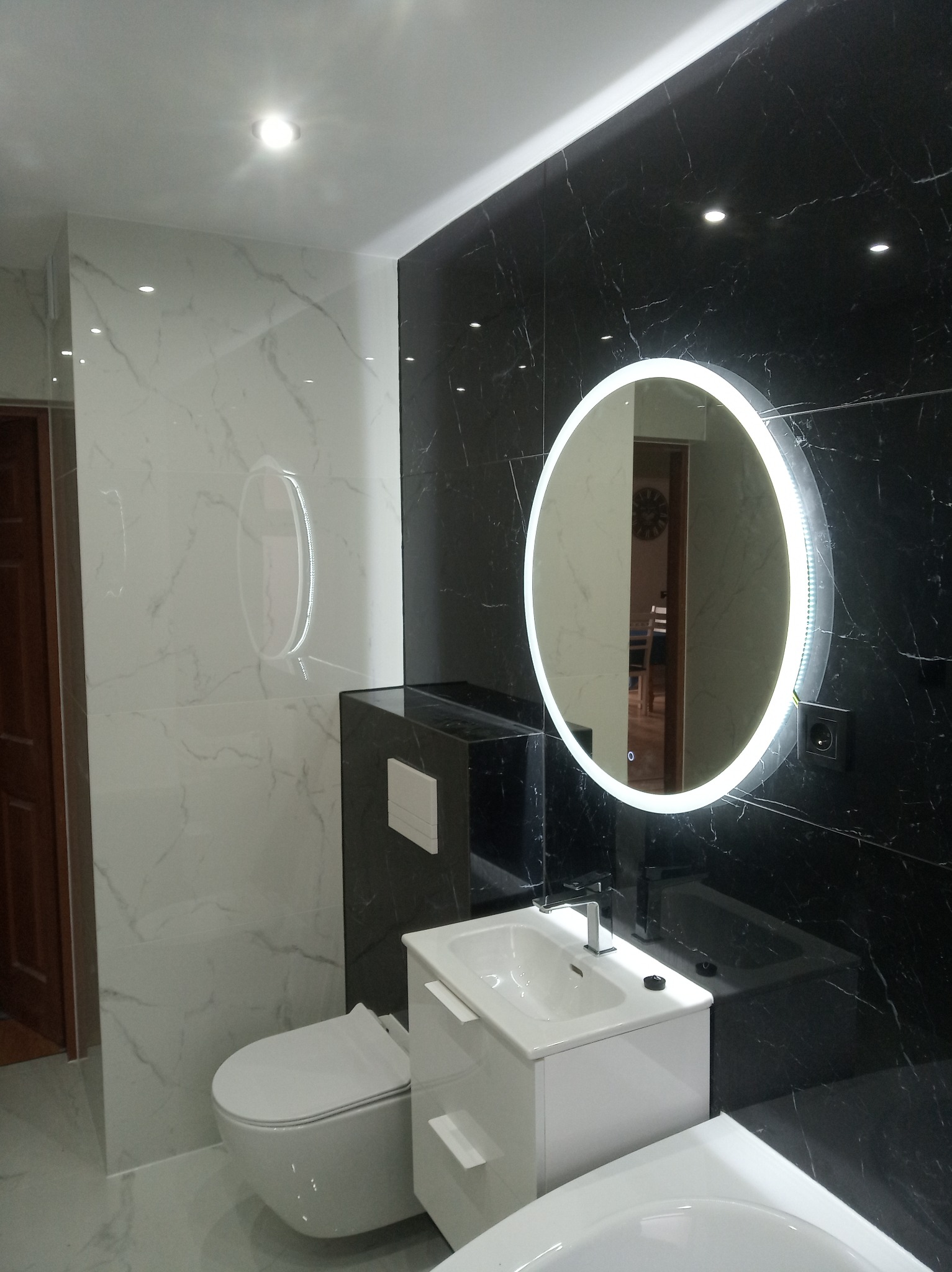 Nowoczesna łazienka z czarno-białymi marmurkowymi płytkami, podwieszaną toaletą, białą szafką z umywalką i okrągłym lustrem z podświetleniem LED. W lustrze odbija się fragment salonu z zegarem...