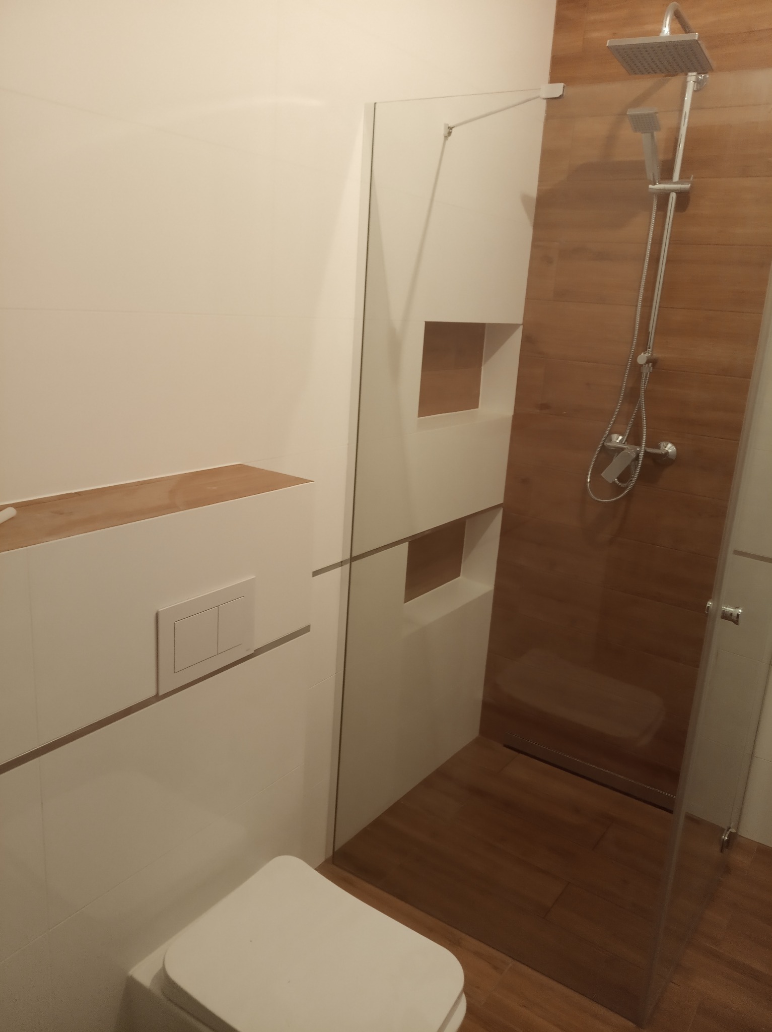 Nowoczesna łazienka z biało-drewnianymi płytkami, kabiną prysznicową typu walk-in z deszczownicą i podtynkową spłuczką WC, w trakcie wykańczania półki nad spłuczką.