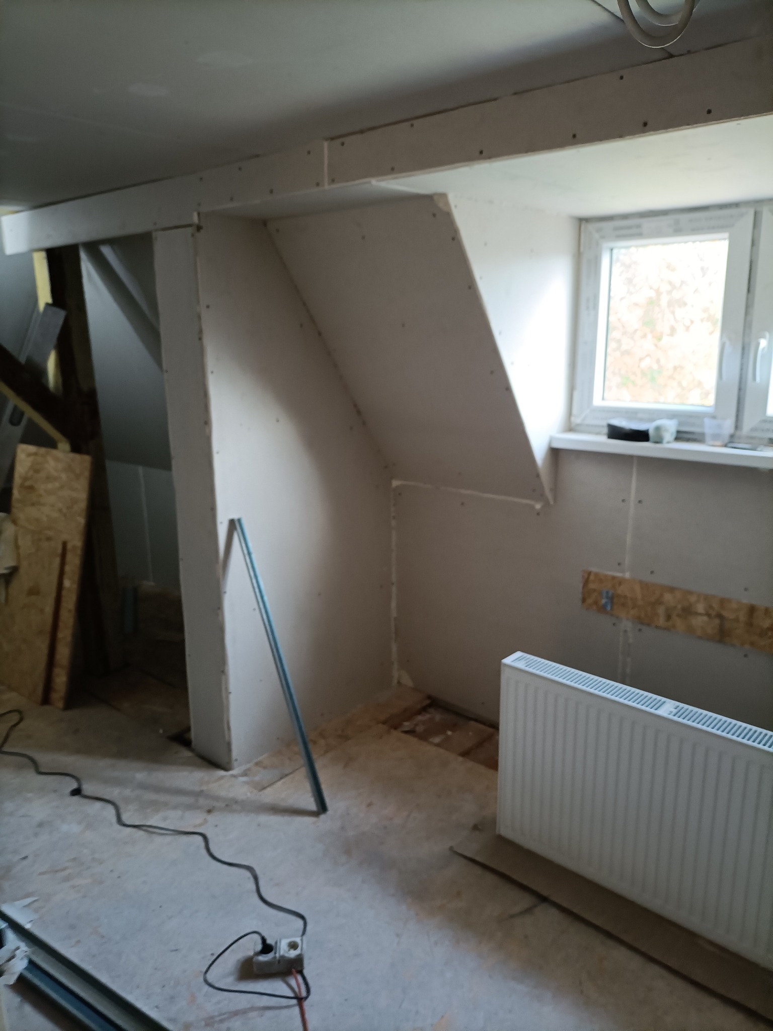 Adaptacja poddasza: ściany i skosy obłożone płytami gipsowo-kartonowymi, widoczny grzejnik, okno dachowe i przewody elektryczne na podłodze.