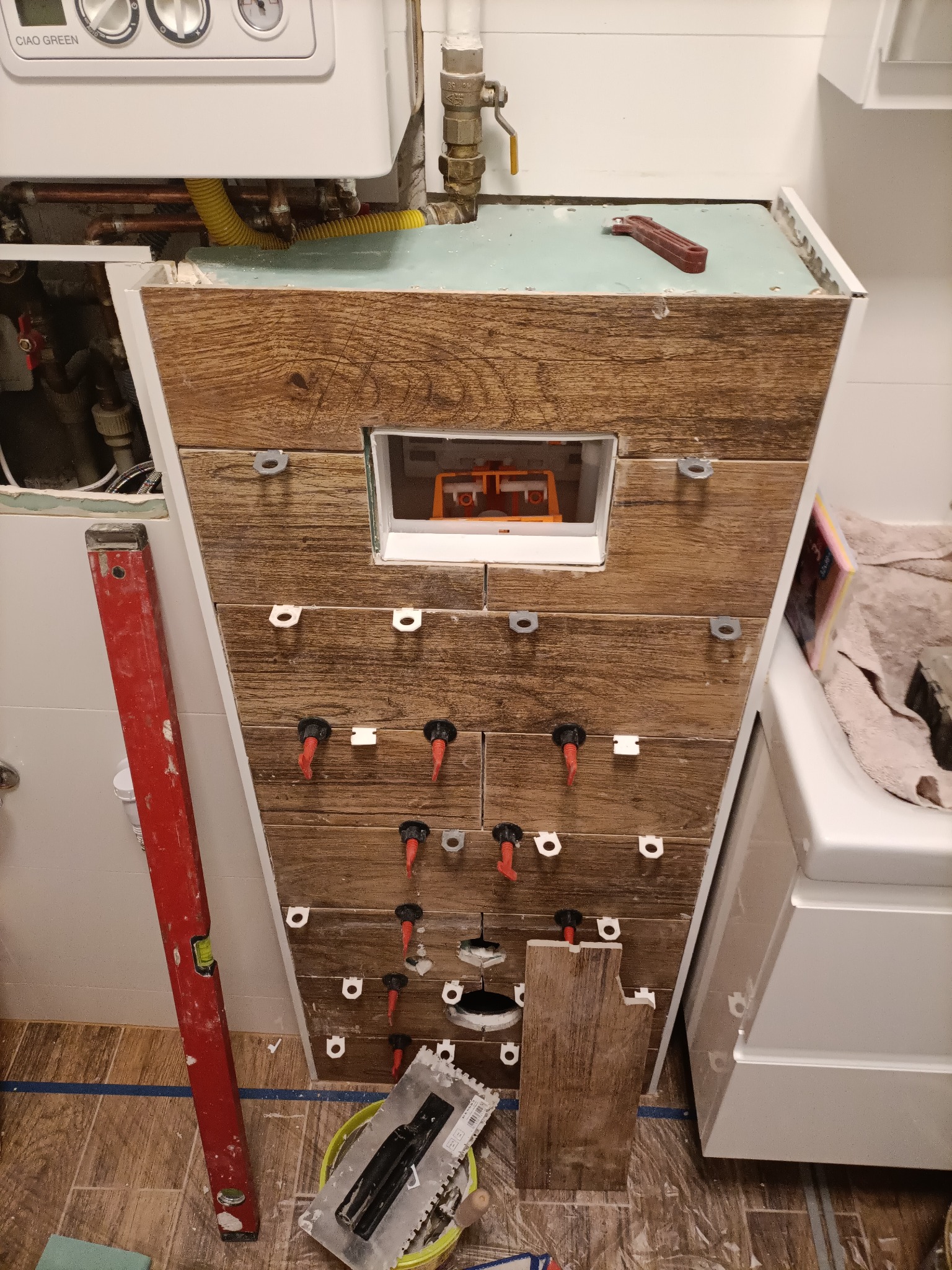 Montaż drewnopodobnych płytek na stelażu podtynkowym WC, widoczne poziomica, wiadro z zaprawą, krzyżyki dystansowe i fragment pieca gazowego