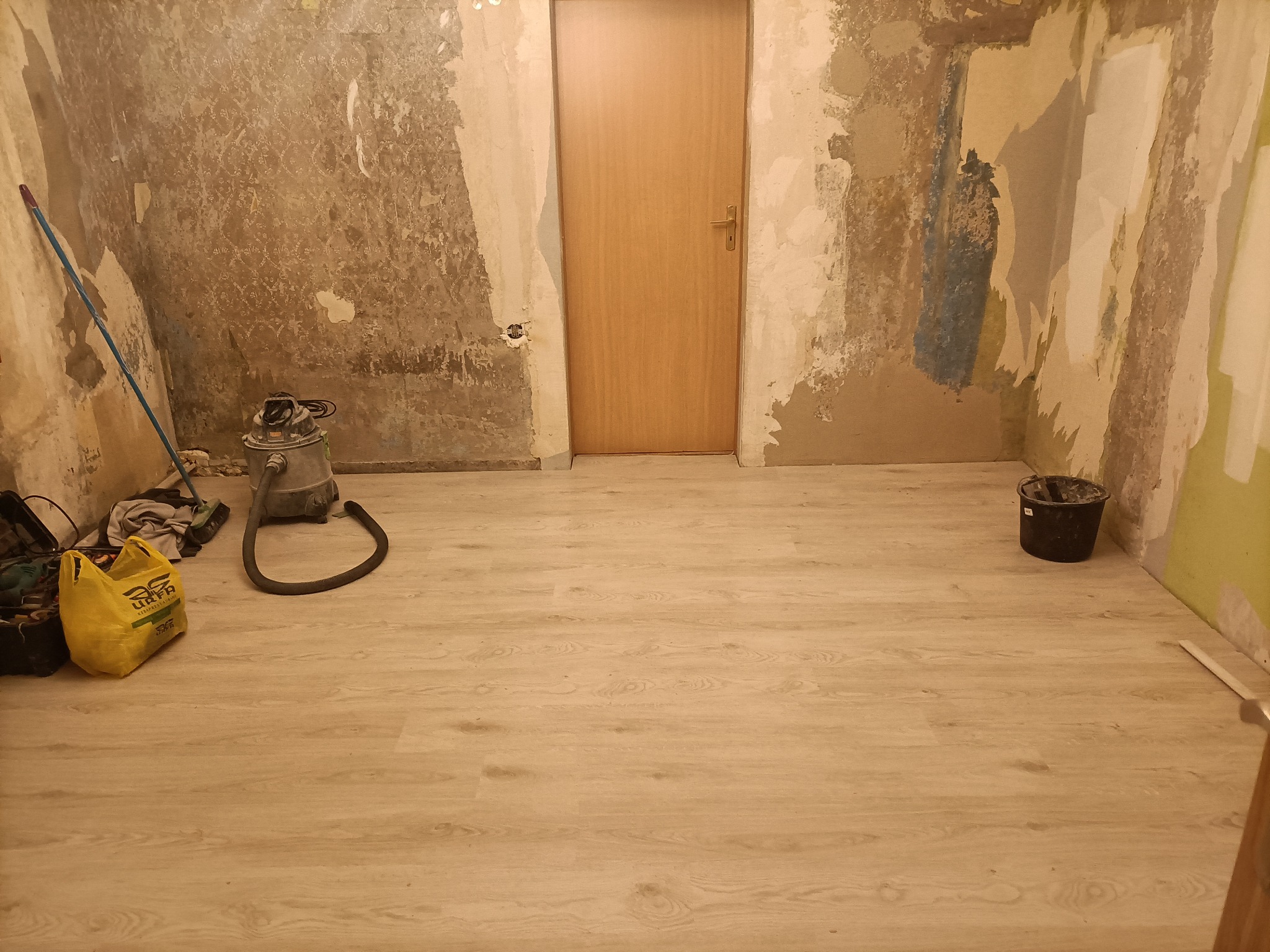 Pokój w trakcie przygotowania do prac wykończeniowych: ściany ze śladami starej farby i tapet, nowa jasna podłoga, widoczny odkurzacz przemysłowy, wiadro i torba z narzędziami.