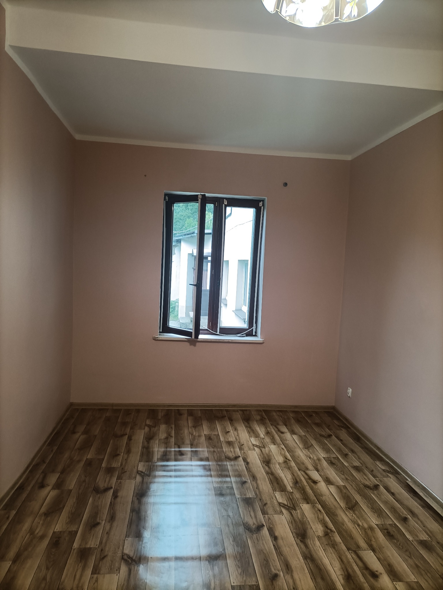 Pusty pokój po remoncie: ściany pomalowane na jasny róż, drewniana podłoga, otwarte okno z widokiem na zieleń.