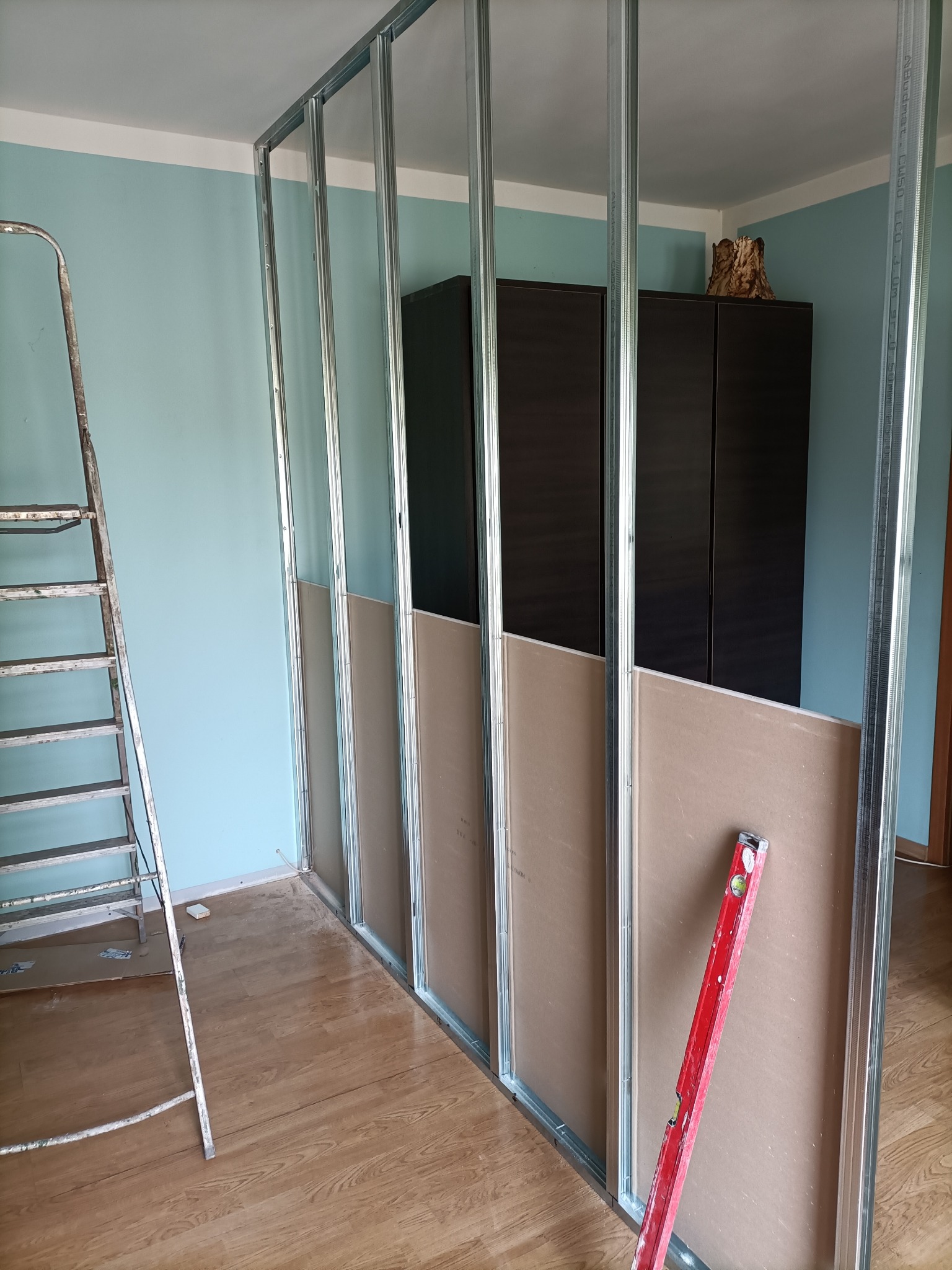 Konstrukcja z profili stalowych i płyt kartonowo-gipsowych w trakcie montażu, poziomica oparta o płytę, drabina przystawiona do ściany na tle błękitnej ściany i ciemnych szafek.