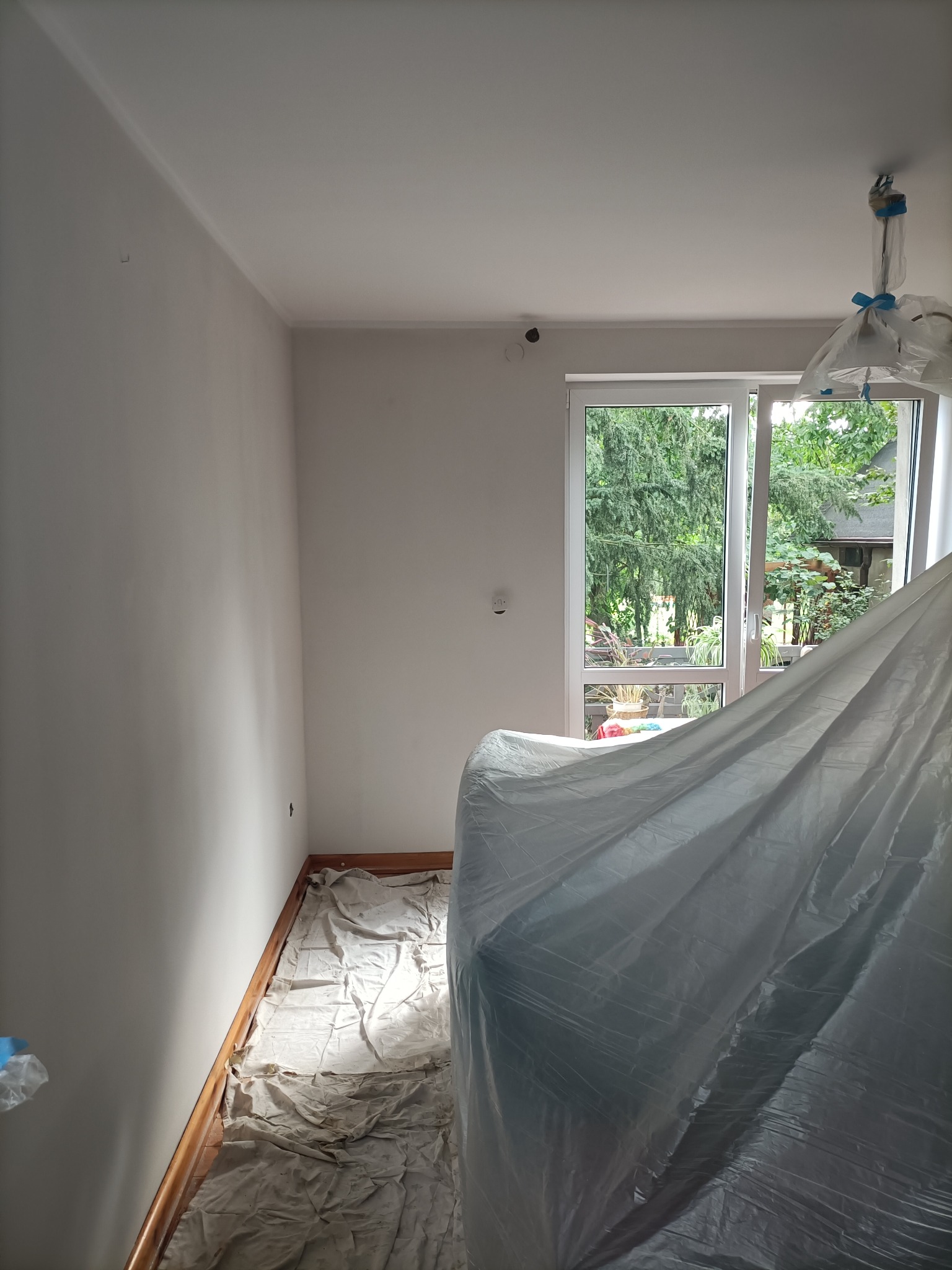 Pokój w trakcie przygotowań do prac wykończeniowych: świeżo pomalowane ściany, podłoga i meble zabezpieczone folią malarską, widok na zieleń za oknem.