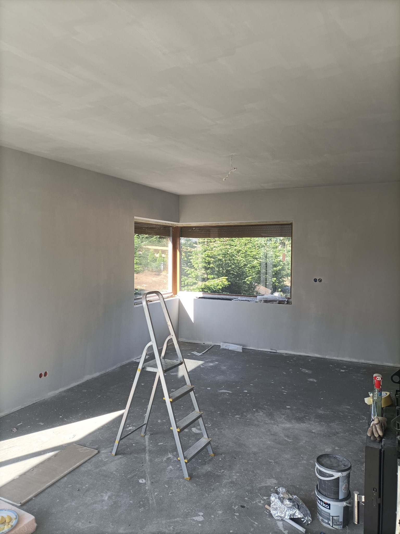 Pokój w trakcie prac wykończeniowych: świeżo położone gładzie na ścianach i suficie, aluminiowa drabina, widok na zieleń przez okno, ślady po pracach na podłodze.