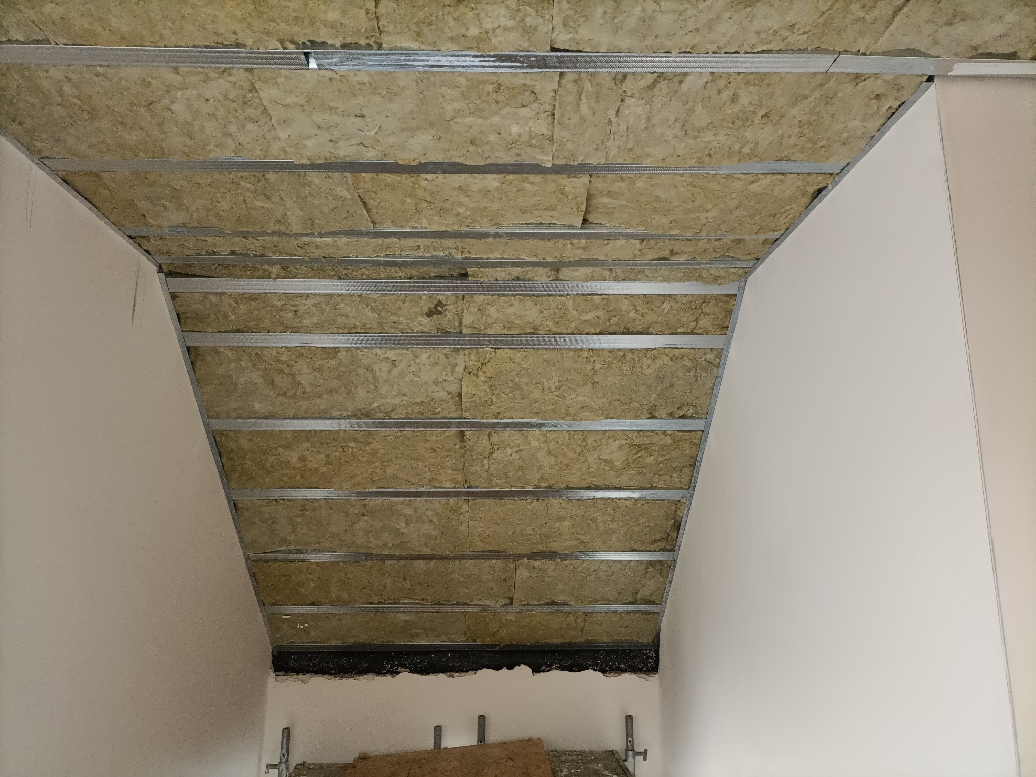 Montaż izolacji z wełny mineralnej w konstrukcji sufitu podwieszanego z metalowych profili, widoczne fragmenty ścian i rusztowania.