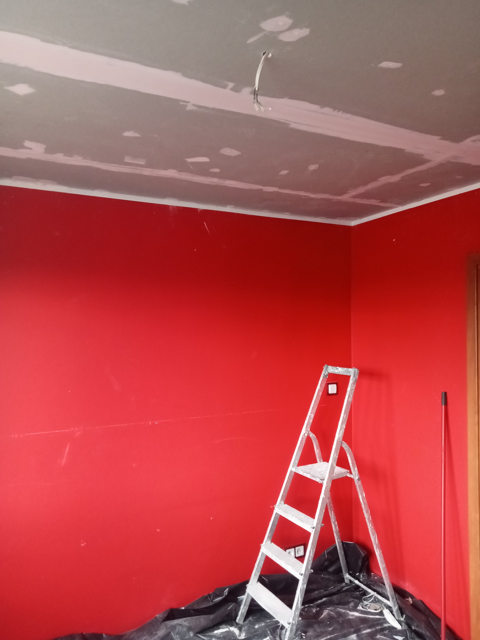 Pokój w trakcie prac: czerwona ściana, częściowo wykończony sufit, drabina malarska i folia ochronna na podłodze.