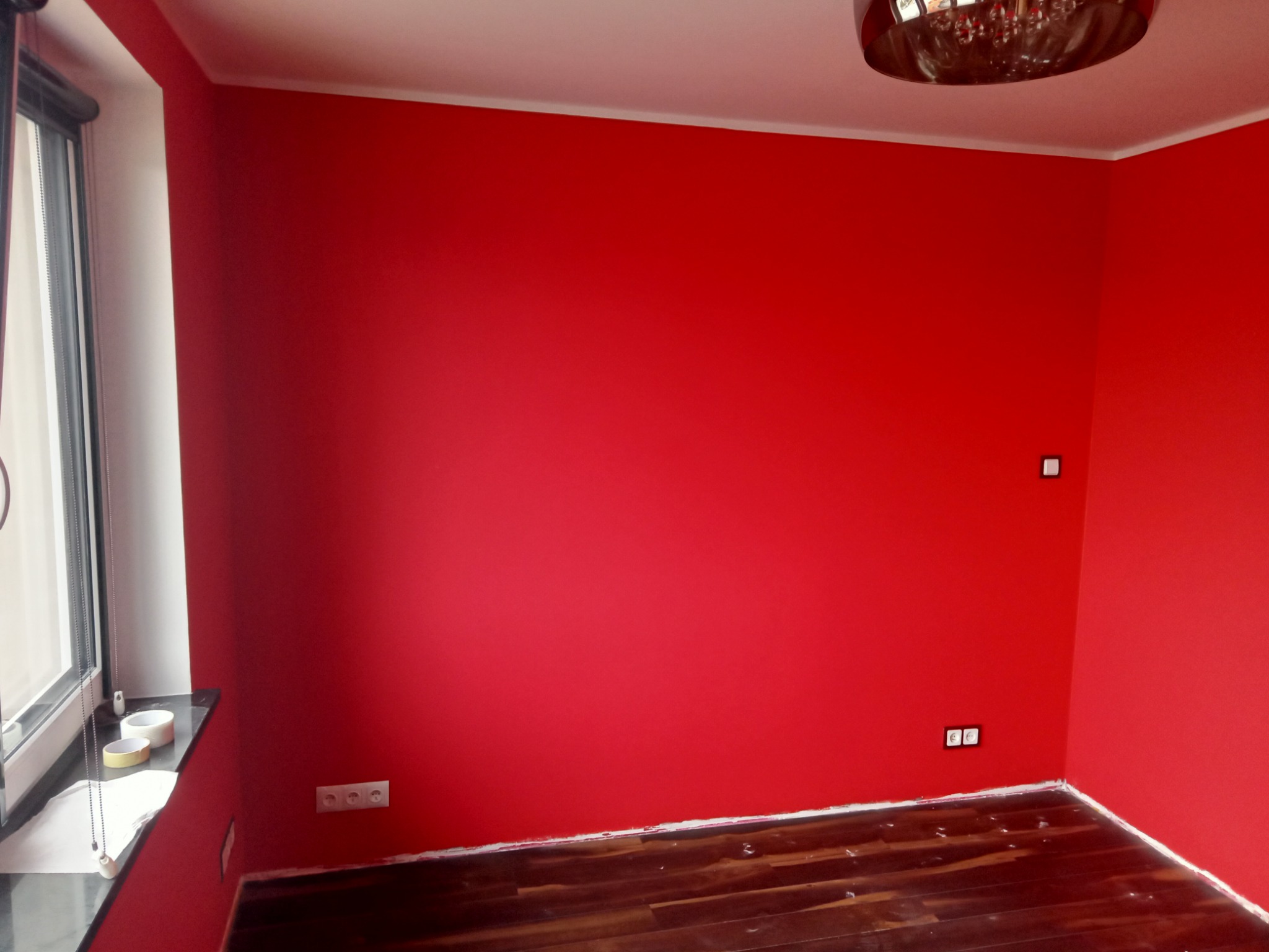 Pomieszczenie po remoncie, ściany pomalowane na intensywny czerwony kolor, ciemna drewniana podłoga, biały sufit z ozdobnym żyrandolem, widoczne gniazdka i włączniki.