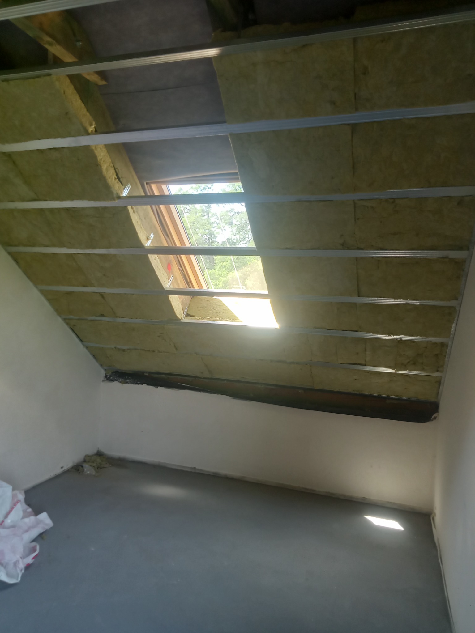 Poddasze z oknem dachowym w trakcie izolacji wełną mineralną, widoczne metalowe profile konstrukcyjne i fragment posadzki z resztkami materiałów budowlanych.