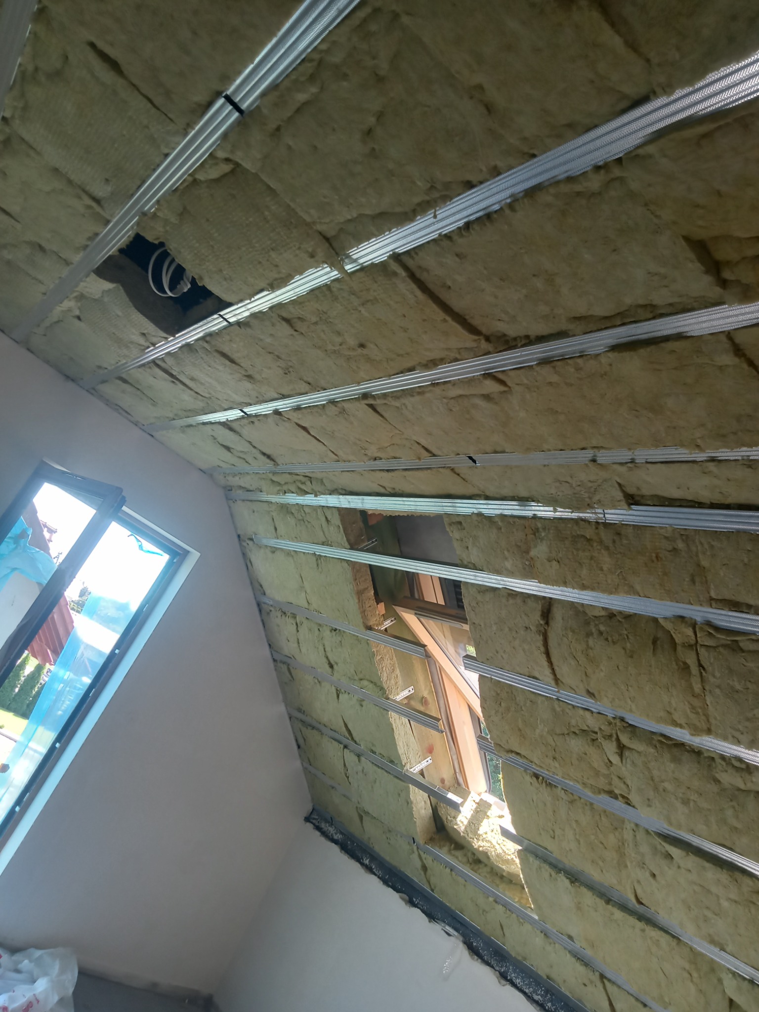 Montaż izolacji z wełny mineralnej na poddaszu, widoczne metalowe profile konstrukcyjne i okno dachowe