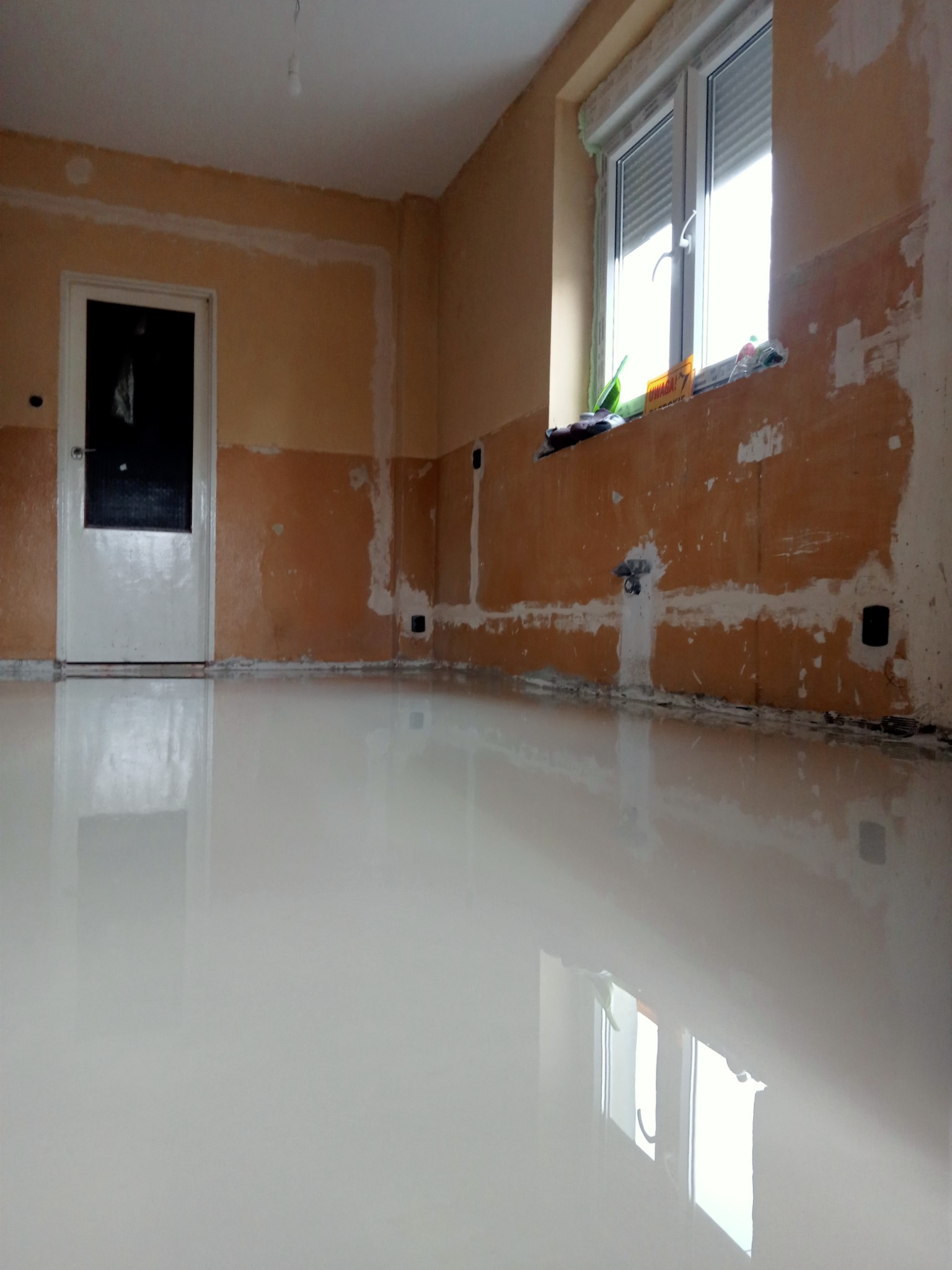 Wnętrze pokoju w trakcie prac przygotowawczych: ściany częściowo pokryte gładzią, świeżo wylana, samopoziomująca wylewka podłogowa, widoczne ślady po instalacjach elektrycznych.