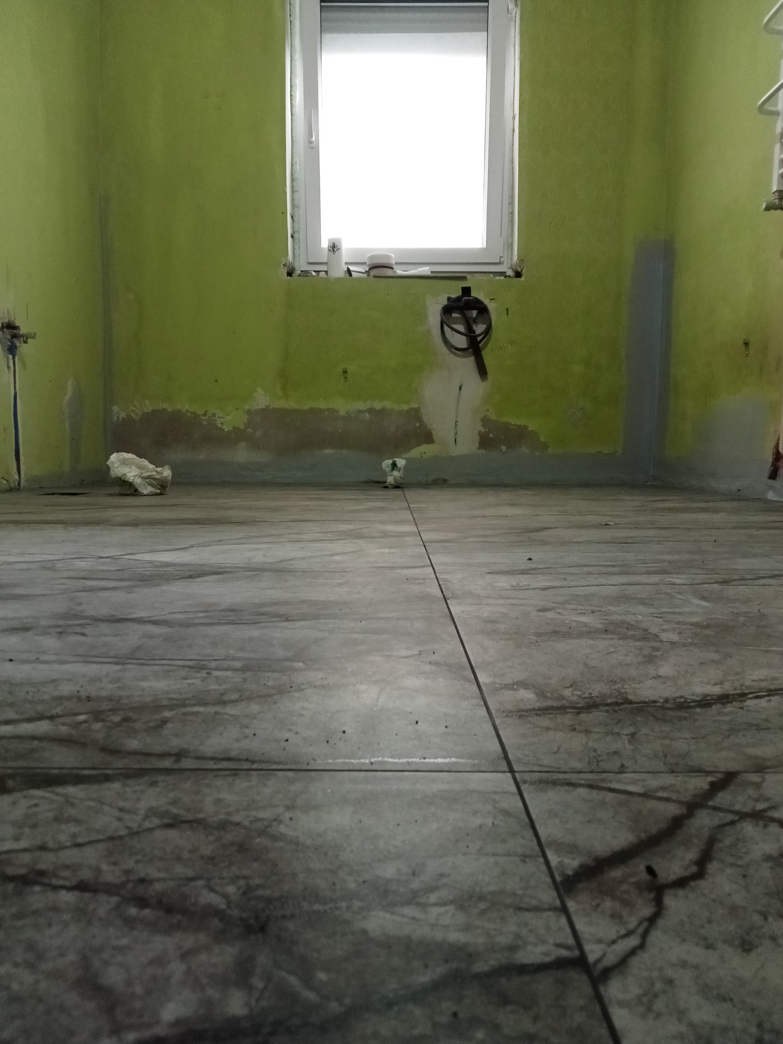 Przygotowane do prac wykończeniowych wnętrze łazienki z zielonymi ścianami, widoczne ślady po instalacjach, surowa podłoga, okno z białymi ramami i resztki materiałów budowlanych.