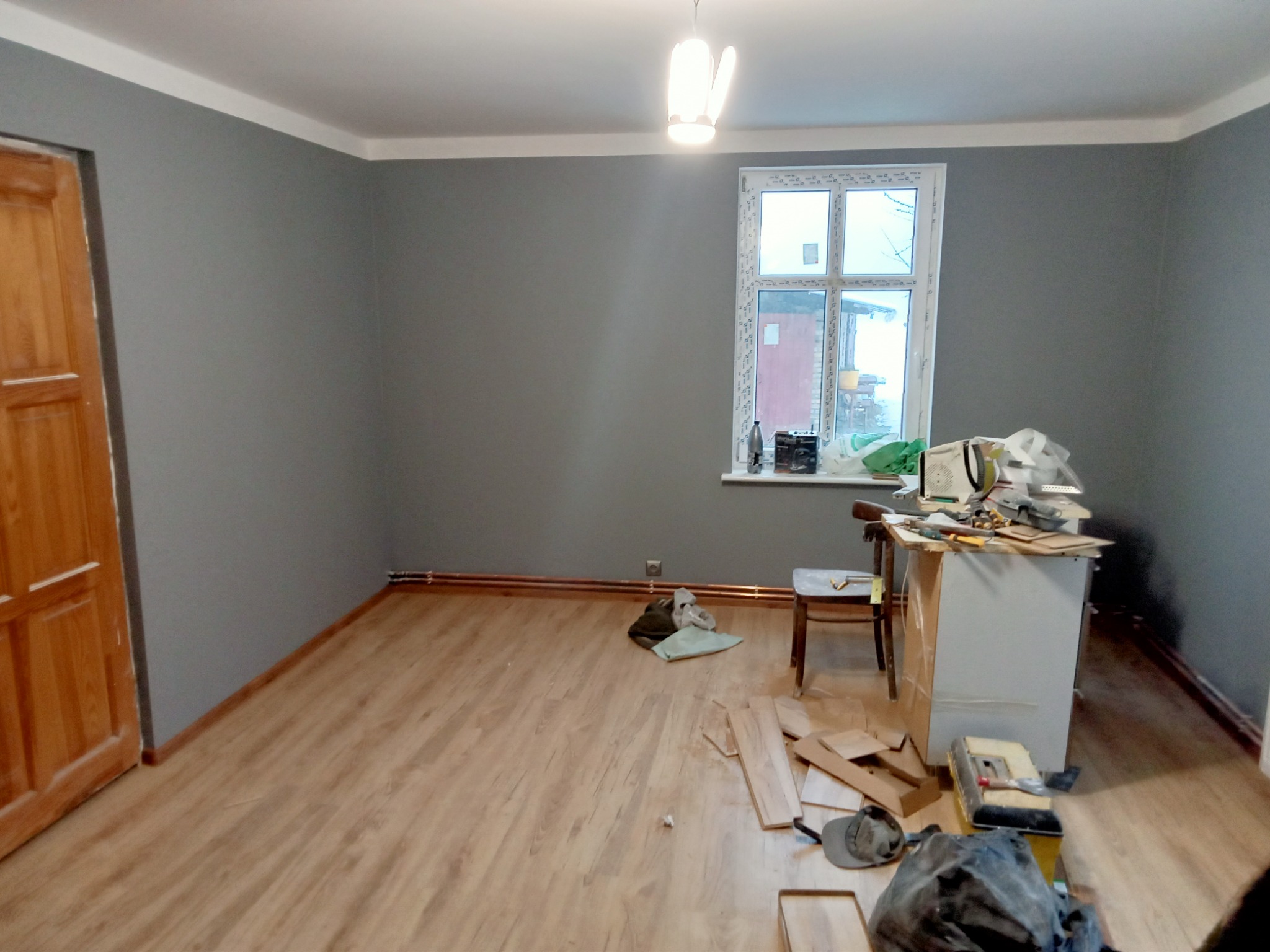 Pokój po malowaniu na szaro z widoczną drewnianą podłogą, białym sufitem i oknem, w trakcie prac wykończeniowych, z narzędziami i materiałami budowlanymi rozłożonymi na podłodze i stole.