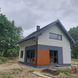 Nowoczesny dom jednorodzinny z szarą dachówką i drewnianą elewacją, częściowo otynkowany, w trakcie wykańczania, z widocznymi materiałami budowlanymi na posesji.