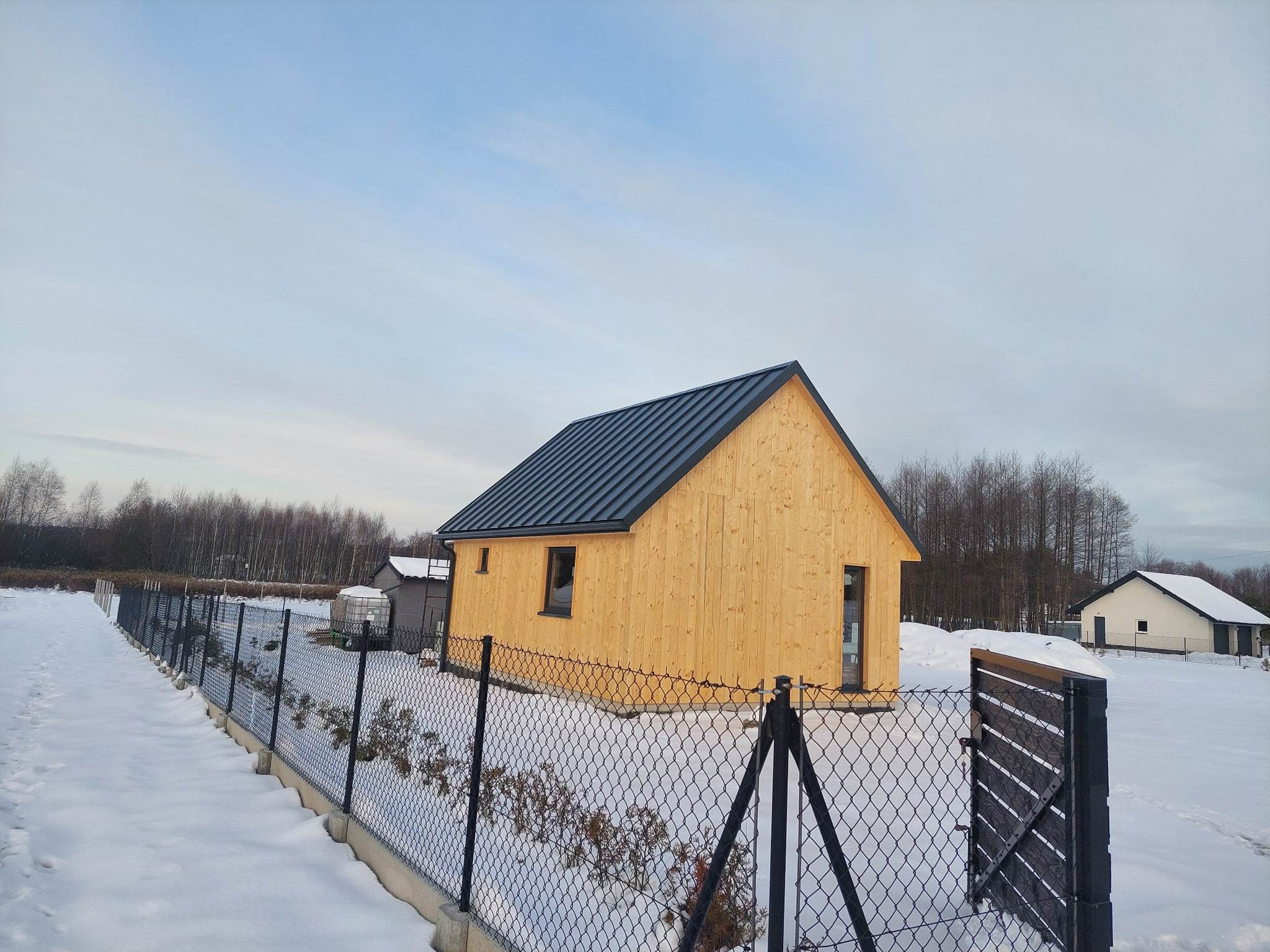 Nowoczesny, parterowy dom z jasnego drewna z ciemnym, metalowym dachem, otoczony siatką ogrodzeniową na betonowej podmurówce, w zimowej scenerii.