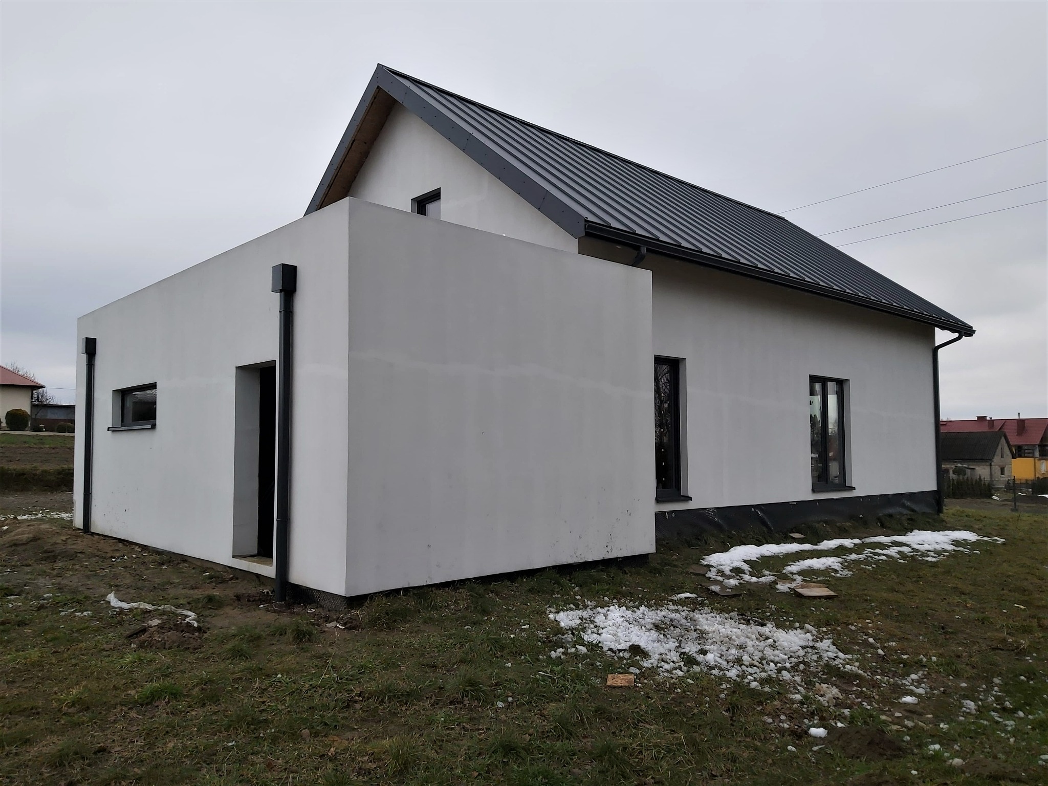 Surowy stan elewacji budynku z dobudowaną, kubistyczną bryłą, widoczne czarne rynny i fragmenty śniegu na trawie.