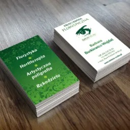 Dwa stosy wizytówek na drewnianym blacie. Jedna sterta z zielonym tłem i napisem 'Florystyka, Hortiterapia, Artystyczna poligrafia, Rękodzieło'. Druga sterta z białym tłem i logo 'Zielone Oczko'...