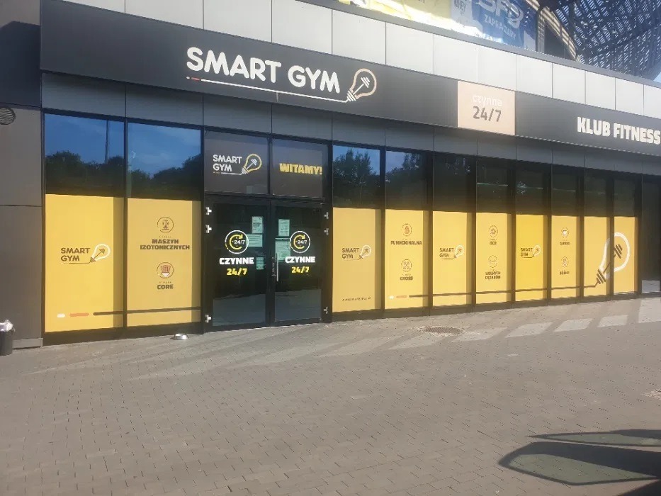 Fasada budynku klubu fitness Smart Gym z dużymi oknami oklejonymi żółtymi foliami z grafikami, logo i informacjami o ofercie, widoczne logo nad wejściem i napis 'Czynne 24/7'.