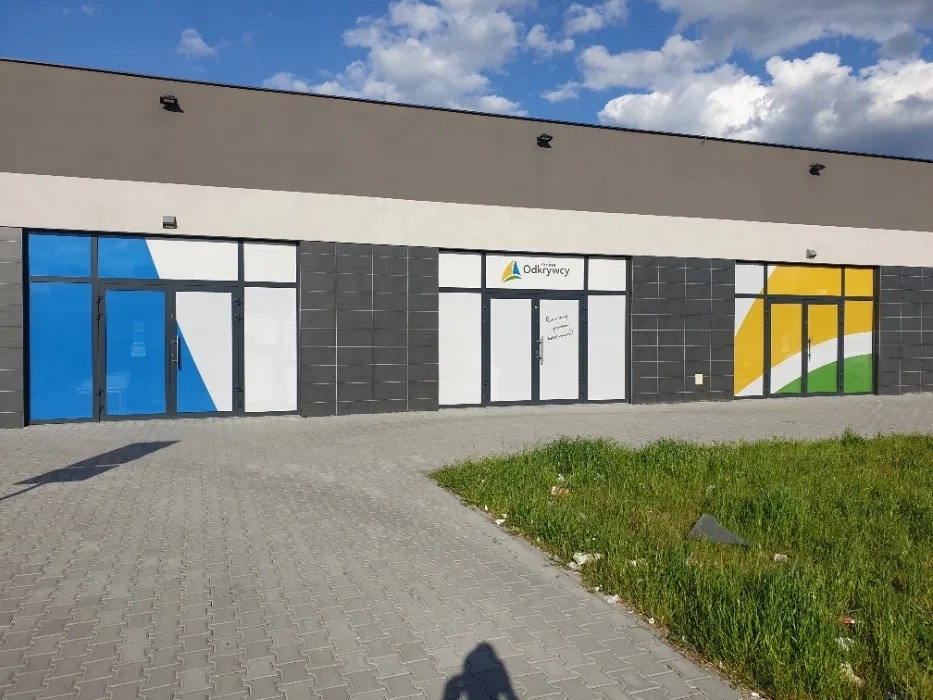 Fasada budynku z trzema wejściami: niebieska folia na szybie po lewej, logo 'Odkrywcy' nad środkowym wejściem, żółto-zielona grafika po prawej. Widoczny chodnik z kostki brukowej i trawa.