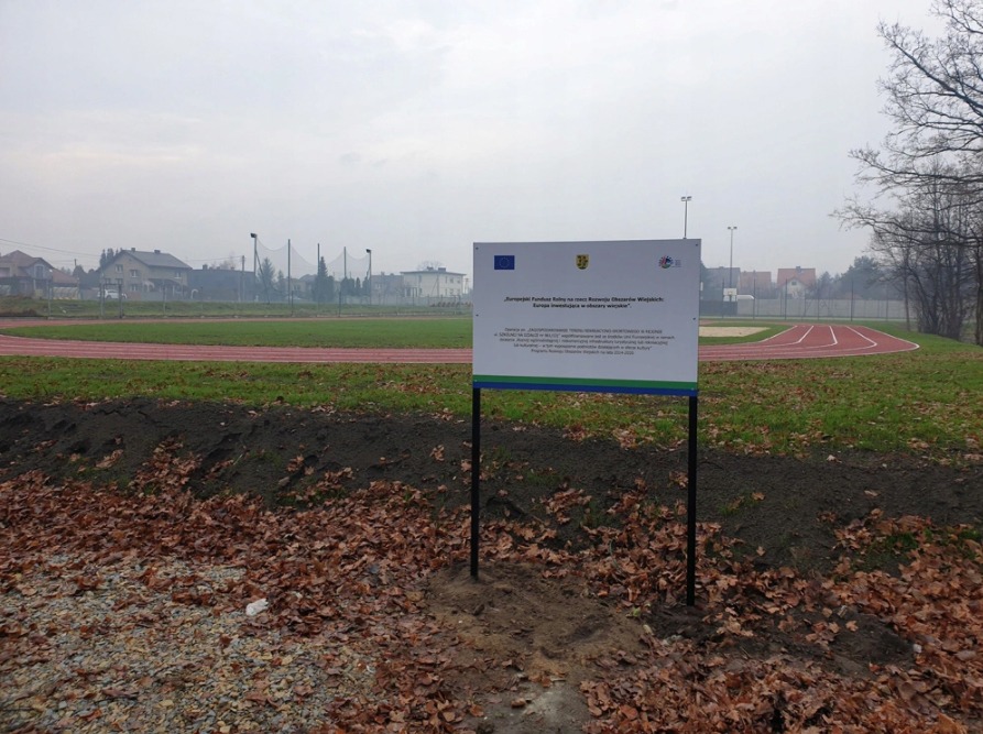 Tablica informacyjna o dofinansowaniu z funduszy europejskich na tle bieżni lekkoatletycznej i zabudowań w pochmurny dzień.