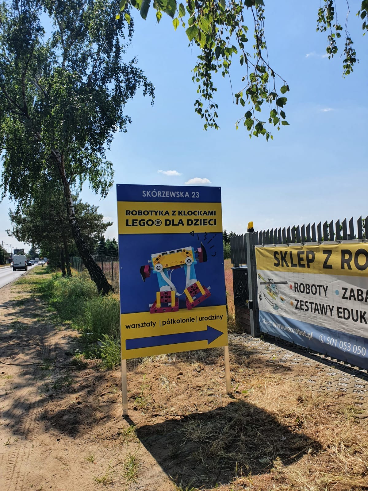 Tablica reklamowa na zewnątrz z ilustracją robota z klocków Lego, informująca o warsztatach robotyki dla dzieci przy ulicy Skórzewskiej 23, obok widoczny baner reklamowy sklepu z robotami.