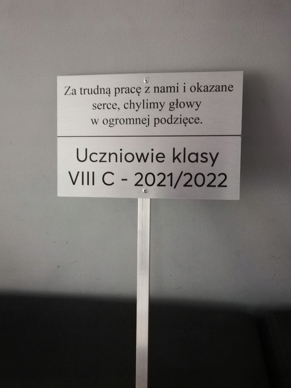 Tabliczka z podziękowaniem od uczniów klasy VIII C z roku 2021/2022, wykonana z metalu w kolorze srebrnym, przymocowana na metalowym słupku.