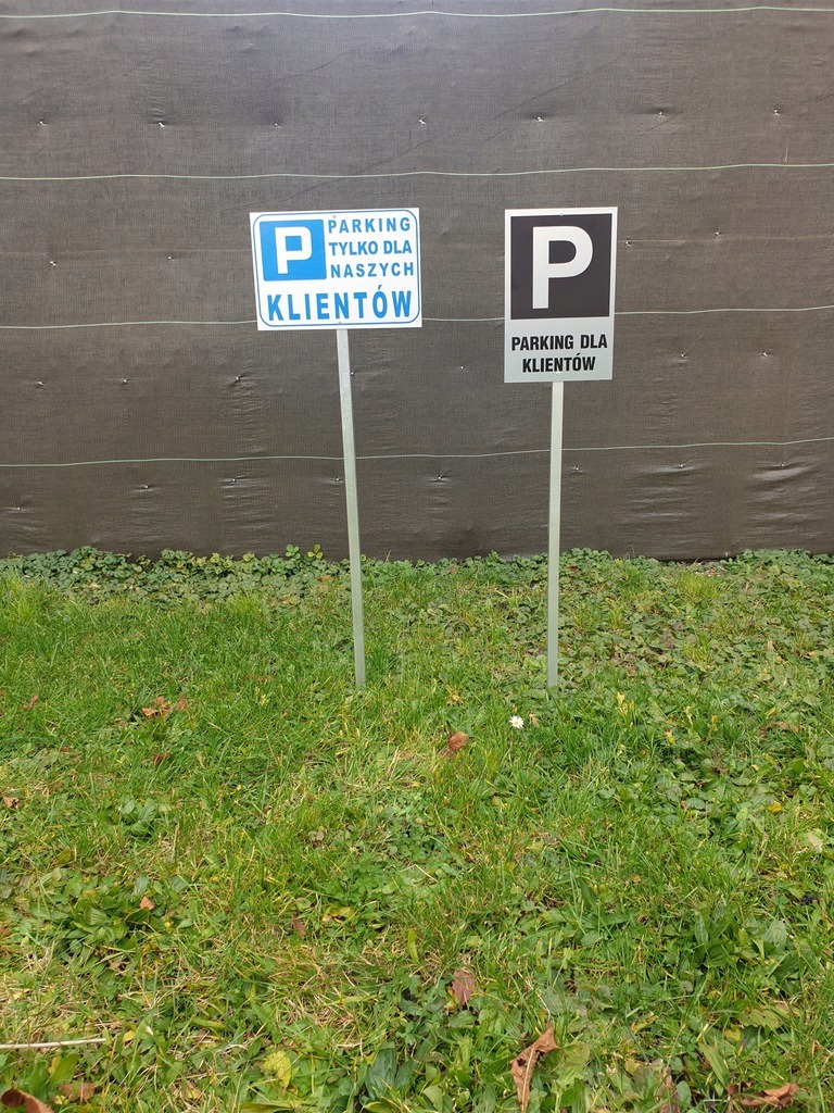 Dwa znaki parkingowe dla klientów na trawniku przed ciemną tkaniną. Jeden znak z niebieskim tłem, drugi z czarnym.