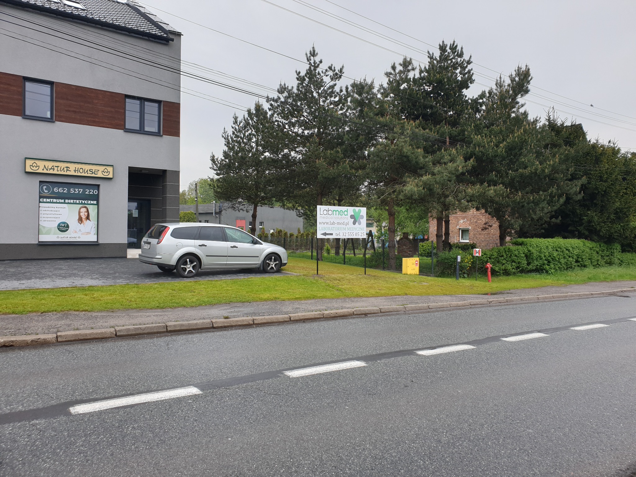 Budynek z szyldem 'Natur House' i tablicą reklamową 'Labmed' na tle drzew, obok zaparkowany srebrny samochód.