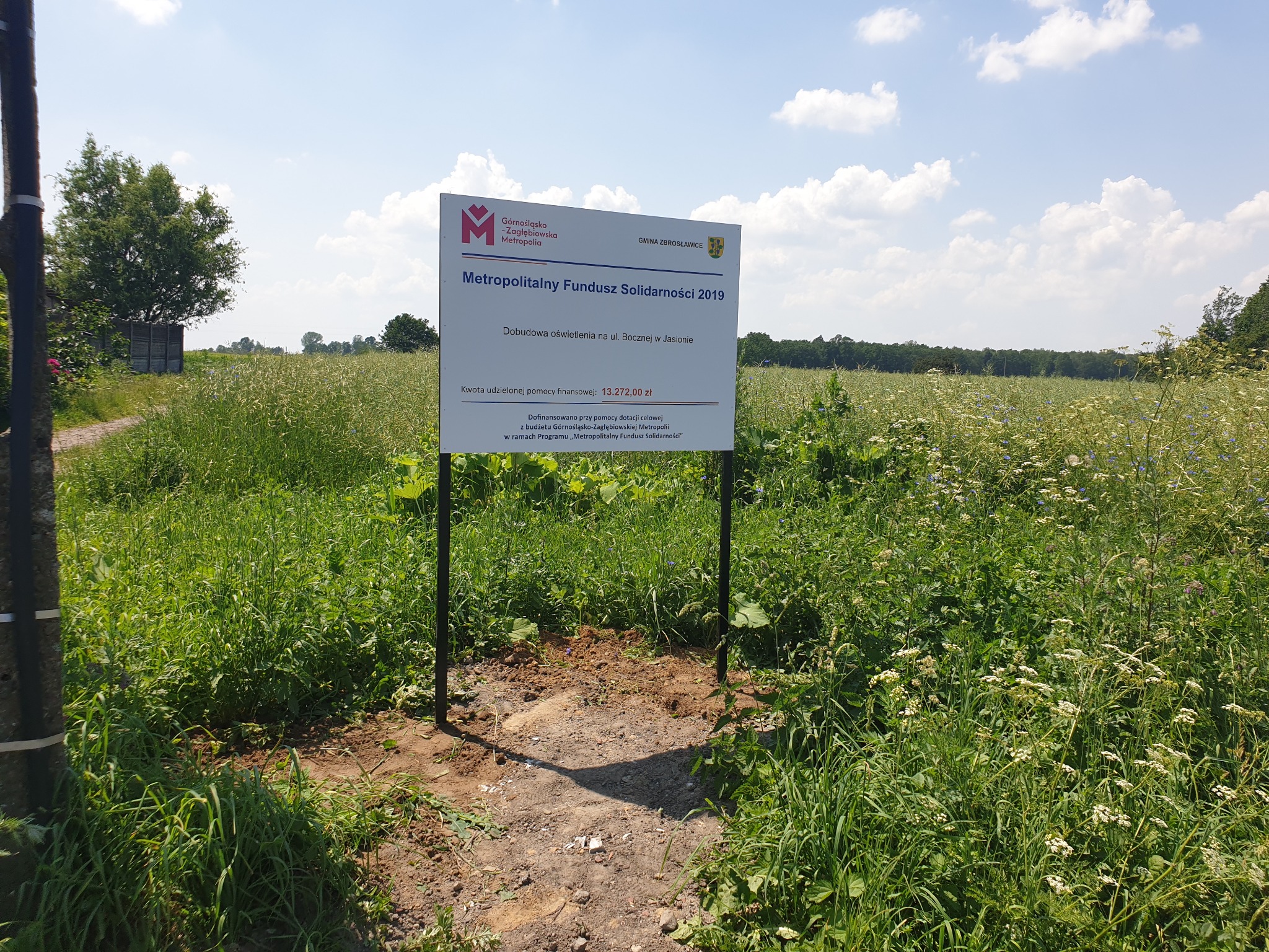 Tablica informacyjna o dofinansowaniu budowy oświetlenia z Metropolitalnego Funduszu Solidarności 2019, umieszczona na tle łąki z kwitnącymi roślinami i błękitnym niebem z chmurami.