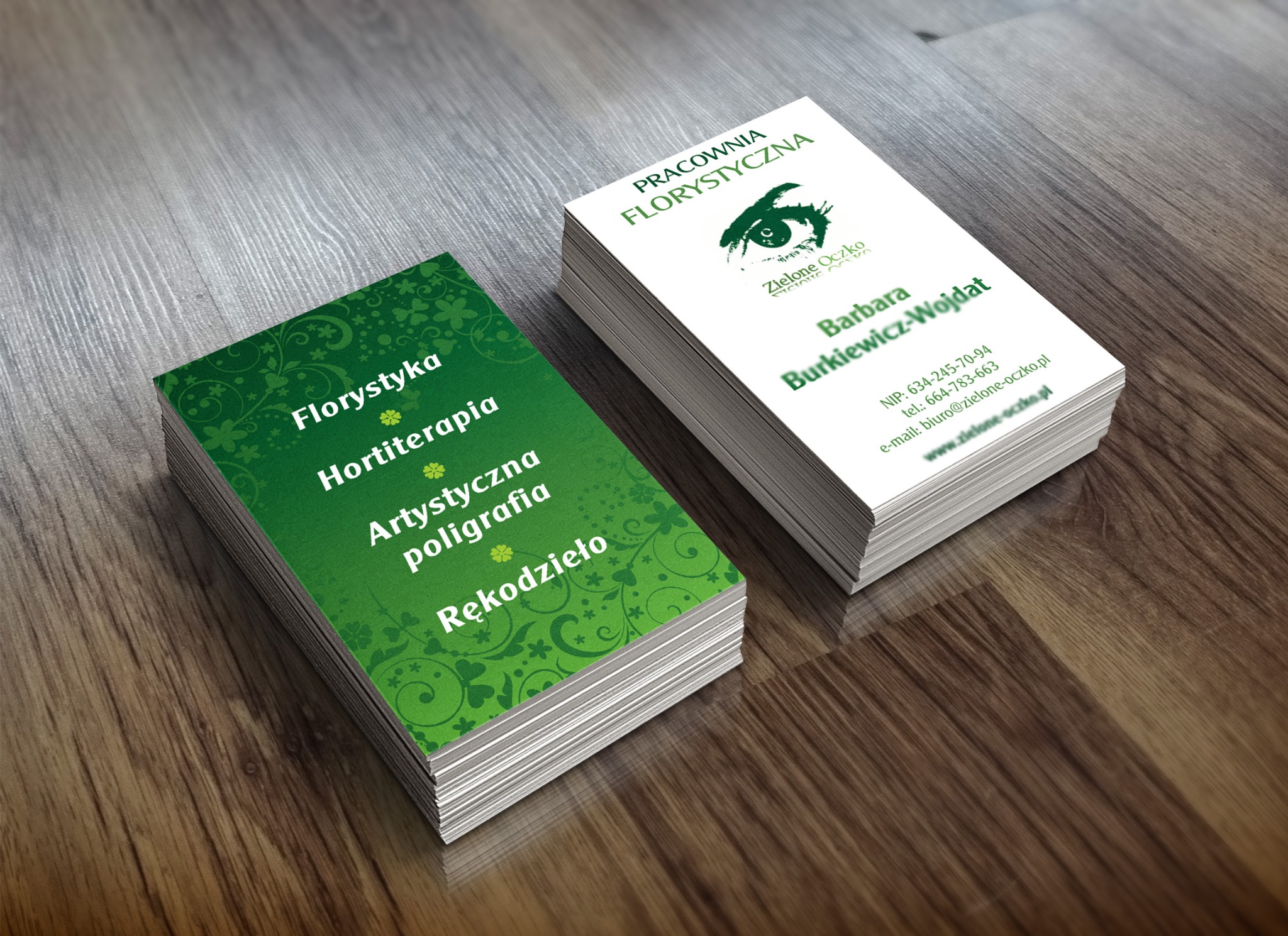 Dwa stosy wizytówek na drewnianym blacie. Jedna sterta z zielonym tłem i napisem 'Florystyka, Hortiterapia, Artystyczna poligrafia, Rękodzieło'. Druga sterta z białym tłem i logo 'Zielone Oczko'...