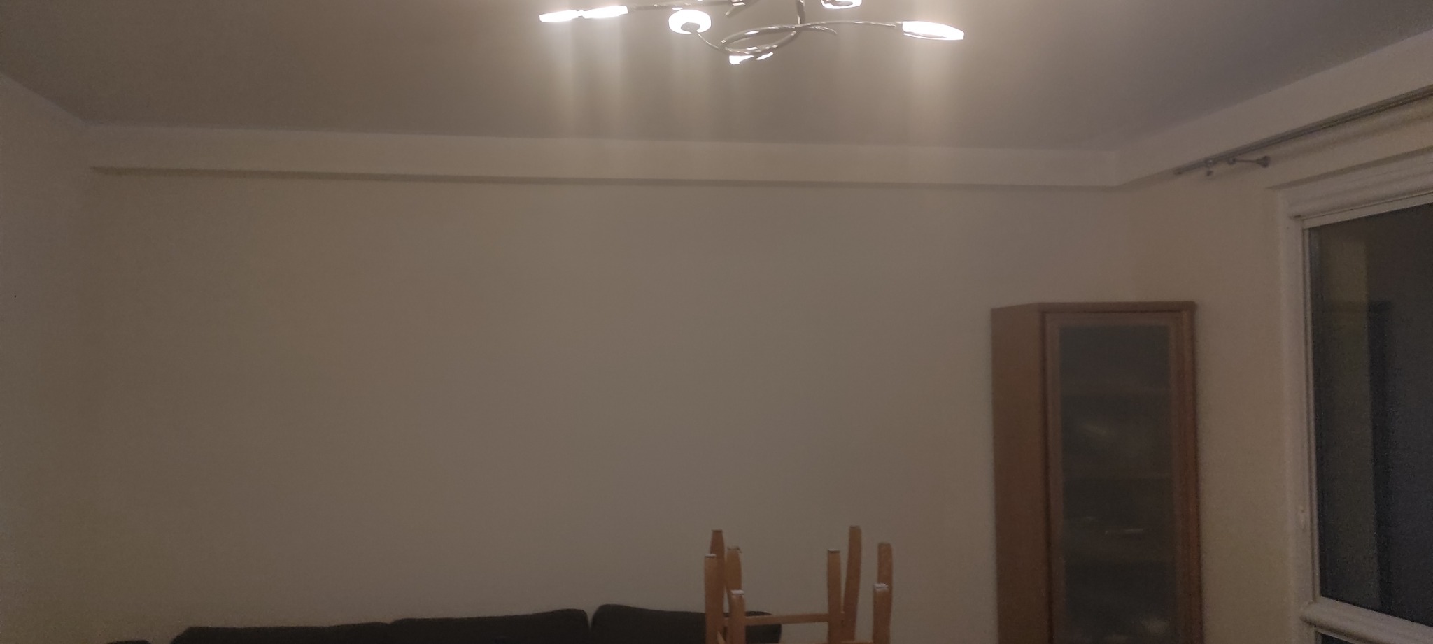 Pusta, beżowa ściana w pokoju z widocznym karniszem, oświetleniem sufitowym, częścią mebla i odwróconym krzesłem, przygotowana do prac wykończeniowych.