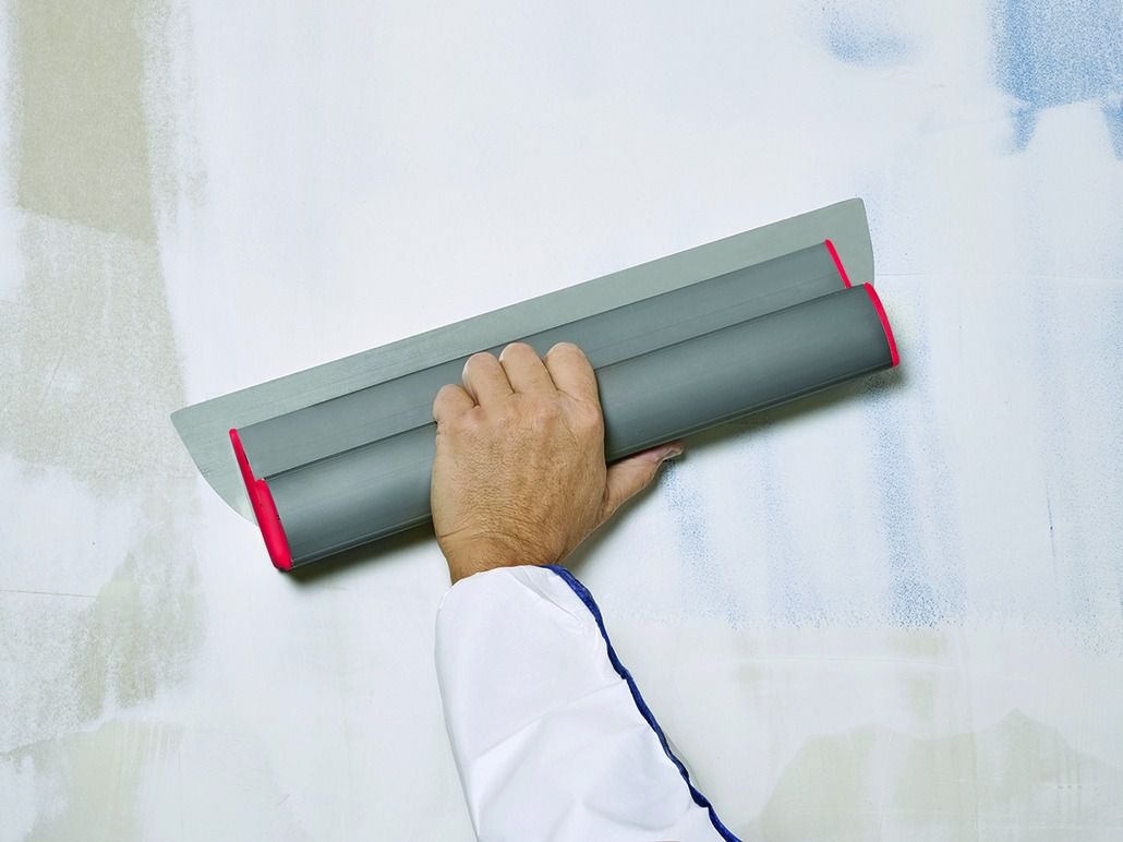 Nakładanie gładzi szpachlowej na ścianę za pomocą dużej, szarej szpachli z czerwonymi elementami, widoczne nierówności podłoża.