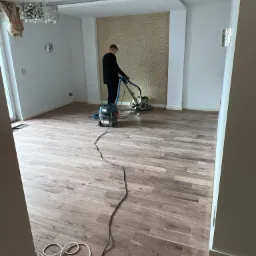 Cyklinowanie parkietu w jasnym pokoju z dekoracyjną ścianą; widoczna maszyna do cyklinowania w użyciu, kabel zasilający na podłodze, oraz nowoczesna lampa sufitowa.