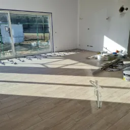 Jasne panele podłogowe ułożone w nowym pomieszczeniu z dużym oknem, widoczne krzyżyki dystansowe, puszki z farbą i narzędzia, w tle przyczepa kempingowa za oknem.