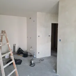 Wnętrze w trakcie prac wykończeniowych: ściany po gładziowaniu, widoczna drabina malarska, wiadro, lampa halogenowa na podłodze i instalacja elektryczna w ścianach.