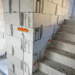 Surowy stan: ściana z betonu komórkowego z pomarańczowymi puszkami elektrycznymi i przewodami, widoczne betonowe schody w tle.