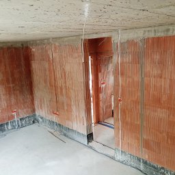 Kamil Chodziuk instalacje elektryczne Limak - Surowy stan: Instalacja elektryczna wewnątrz budynku z czerwonej cegły. Widoczne kable i puszki instalacyjne. Podłoga betonowa, sufit surowy. W tle otwór drzwiowy.