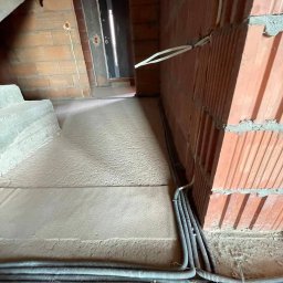 Kamil Chodziuk instalacje elektryczne Limak - Kable elektryczne w peszlach ułożone wzdłuż ściany z czerwonej cegły w surowym wnętrzu budynku w trakcie budowy. Widoczne puszki elektryczne w ścianie.