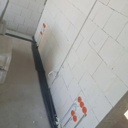 Kamil Chodziuk instalacje elektryczne Limak - Ściana z instalacją elektryczną w trakcie montażu: kable w peszlach prowadzone wzdłuż ściany, puszki elektryczne z pomarańczowymi dekielkami. Surowy stan.