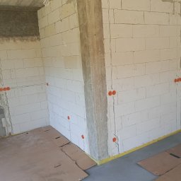 Kamil Chodziuk instalacje elektryczne Limak - Wnętrze w stanie surowym z widoczną instalacją elektryczną w ścianach z betonu komórkowego. Pomarańczowe puszki elektryczne rozmieszczone wzdłuż ścian. Podłoga zabezpieczona kartonem.