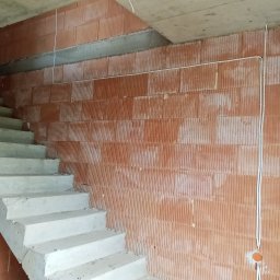 Kamil Chodziuk instalacje elektryczne Limak - Surowa ściana z cegły dziurawki z poprowadzoną instalacją elektryczną w peszlu, widoczne betonowe schody w surowym stanie. Budowa domu w trakcie realizacji.