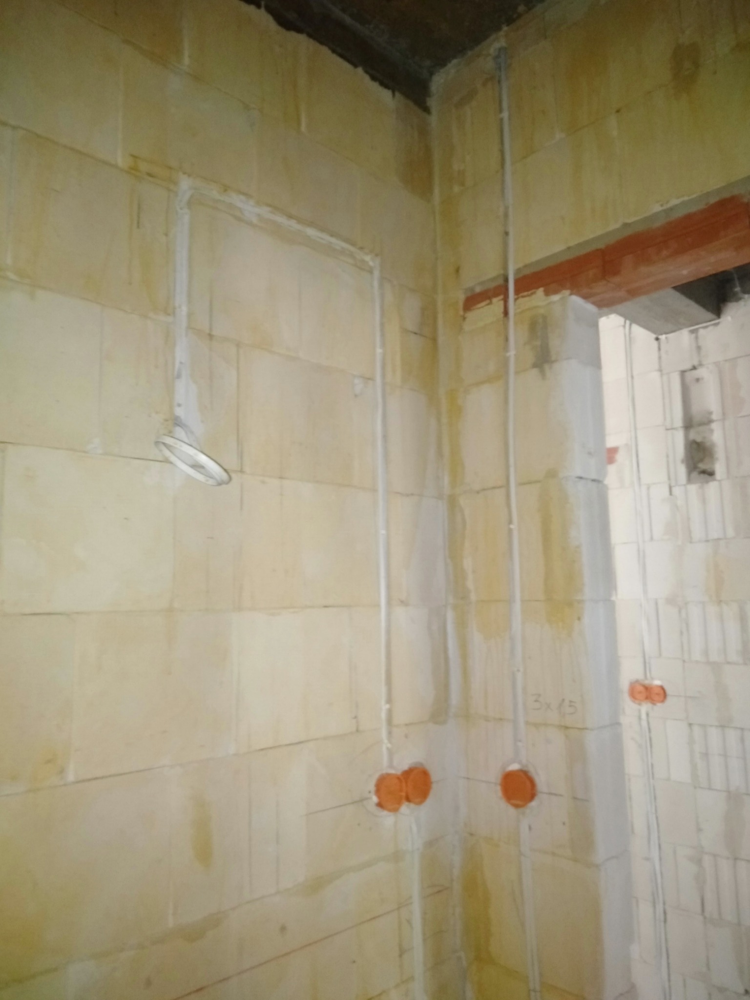 Surowe ściany z betonu komórkowego w trakcie instalacji elektrycznej: widoczne peszle, puszki instalacyjne oraz fragment oprawy oświetleniowej z kablem.